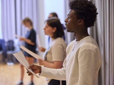 A row of people stand in a rehearsal room holding scripts, practising their speeches